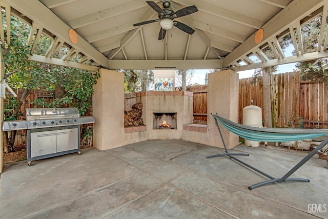 view of patio with a warm lit fireplace, ceiling fan, a grill, fence, and a gazebo