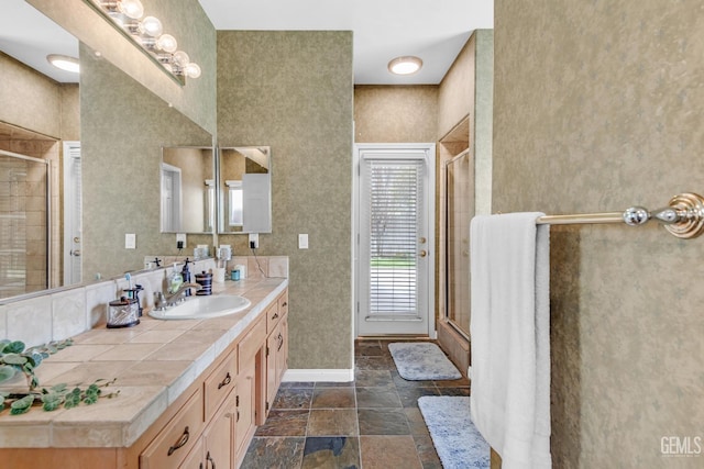 full bathroom with a shower with door, stone tile floors, baseboards, and vanity