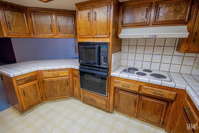 kitchen featuring black appliances, tile counters, ventilation hood, and brown cabinetry