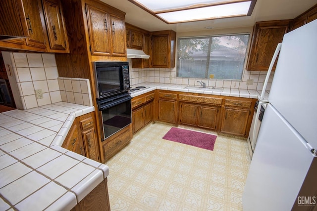 kitchen with tile countertops, light floors, a sink, under cabinet range hood, and black appliances