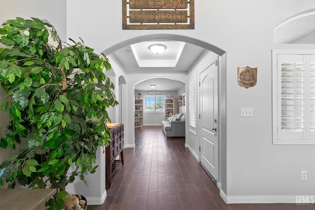 hallway featuring baseboards, a tray ceiling, arched walkways, and dark wood-type flooring