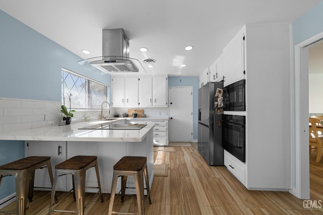 kitchen featuring a peninsula, black appliances, light wood finished floors, island exhaust hood, and tasteful backsplash