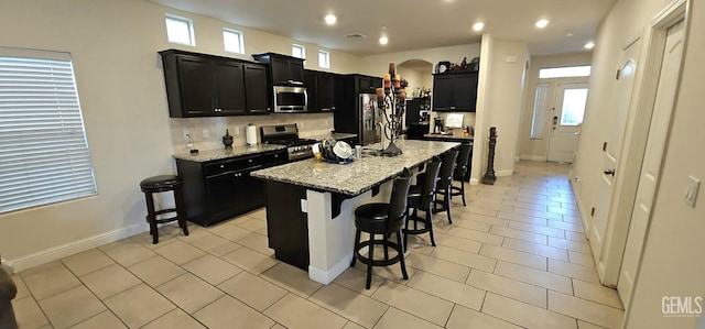 kitchen with a breakfast bar area, light stone counters, light tile patterned floors, appliances with stainless steel finishes, and an island with sink