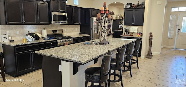 kitchen featuring a kitchen island with sink, stainless steel appliances, tasteful backsplash, light stone countertops, and a kitchen bar