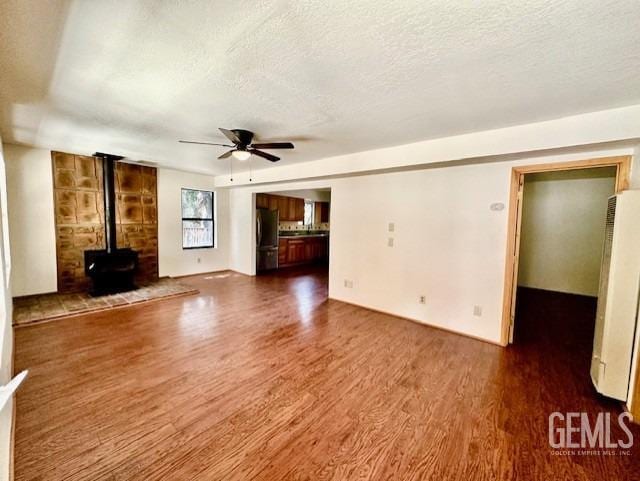unfurnished living room featuring a wood stove, a textured ceiling, a ceiling fan, and wood finished floors