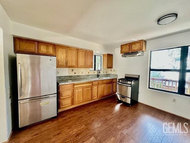 kitchen featuring brown cabinetry, appliances with stainless steel finishes, dark wood-style flooring, a sink, and under cabinet range hood