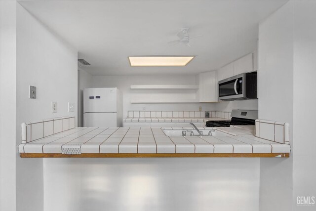 kitchen with tile countertops, stove, ceiling fan, white fridge, and white cabinetry