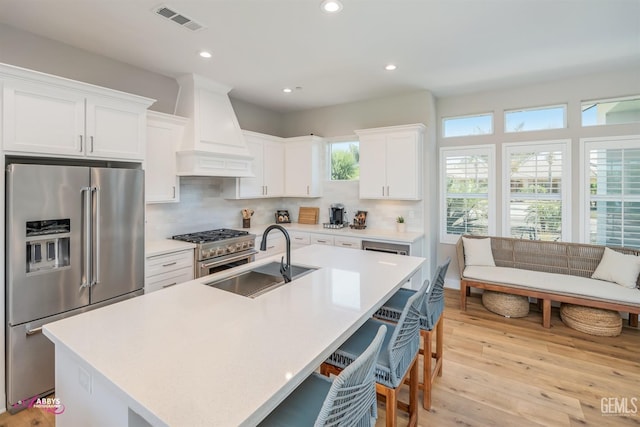 kitchen with custom exhaust hood, high end appliances, a kitchen island with sink, sink, and white cabinetry