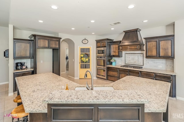 kitchen featuring custom exhaust hood, backsplash, a large island with sink, sink, and stainless steel appliances