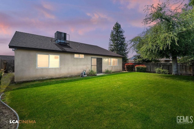 back of property at dusk with a yard, a shingled roof, stucco siding, and fence