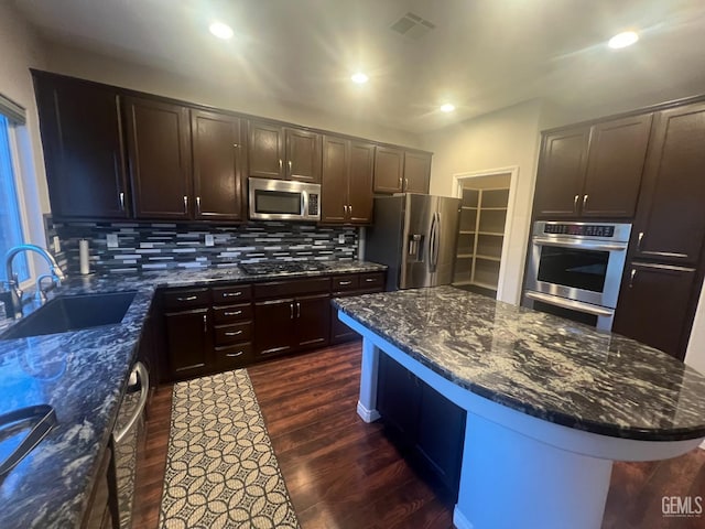 kitchen featuring stainless steel appliances, a sink, backsplash, dark wood-style floors, and dark stone countertops