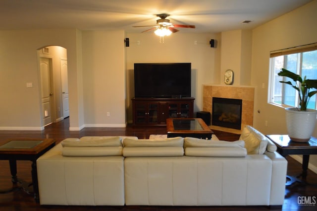 living area featuring arched walkways, a tiled fireplace, a ceiling fan, wood finished floors, and baseboards