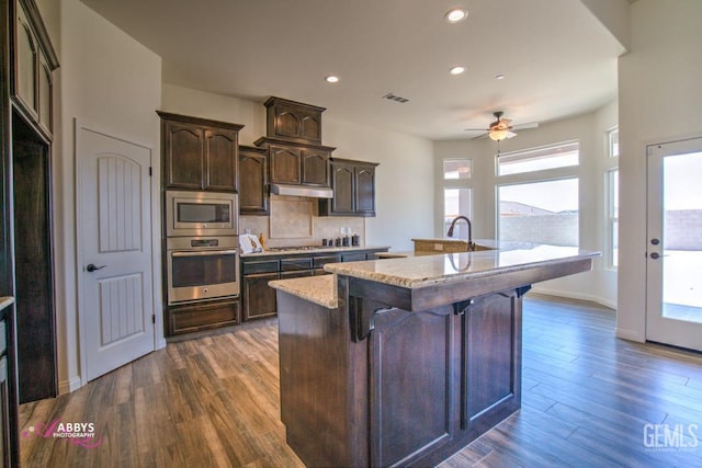 kitchen featuring backsplash, dark hardwood / wood-style flooring, stainless steel appliances, dark brown cabinets, and a center island with sink