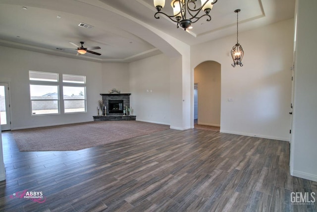 unfurnished living room featuring dark wood-type flooring, a tray ceiling, ceiling fan with notable chandelier, and a stone fireplace