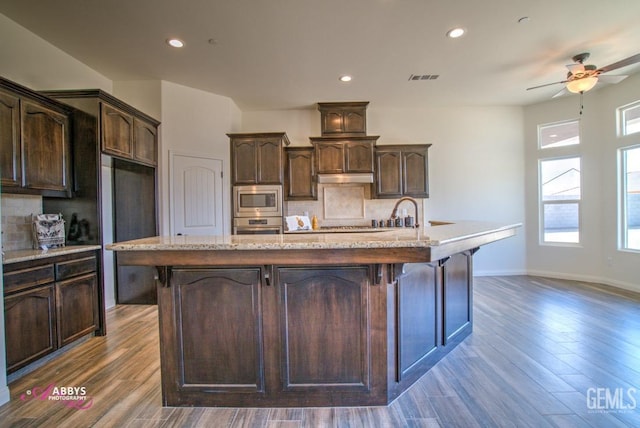 kitchen with appliances with stainless steel finishes, dark hardwood / wood-style flooring, decorative backsplash, dark brown cabinets, and a center island with sink