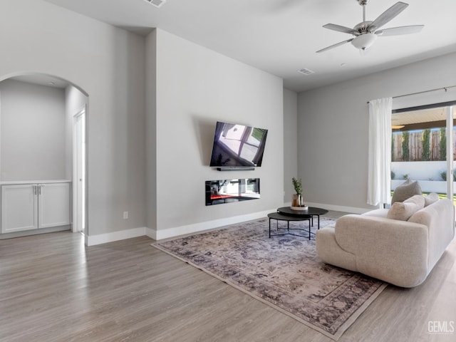 living area featuring arched walkways, a ceiling fan, baseboards, light wood-type flooring, and a glass covered fireplace