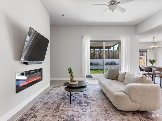 living area featuring visible vents, a glass covered fireplace, wood finished floors, baseboards, and ceiling fan with notable chandelier