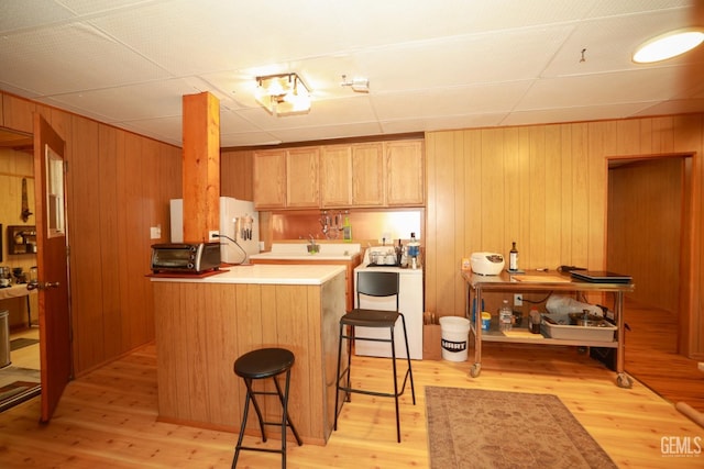 kitchen featuring a drop ceiling, wooden walls, light hardwood / wood-style floors, a kitchen bar, and kitchen peninsula