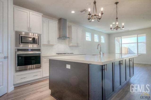 kitchen with wall chimney exhaust hood, a notable chandelier, a center island with sink, white cabinets, and appliances with stainless steel finishes