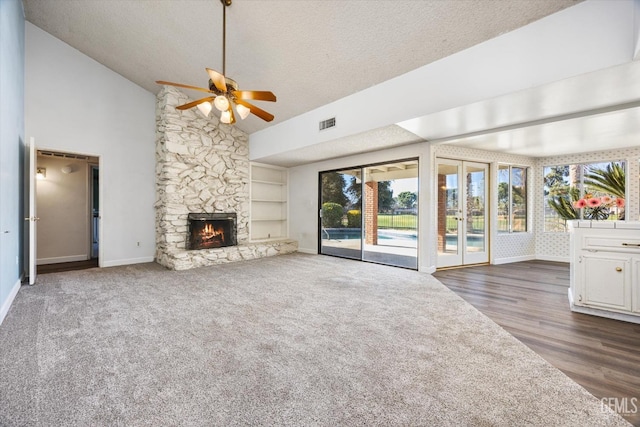 unfurnished living room with a textured ceiling, dark colored carpet, built in features, ceiling fan, and a stone fireplace