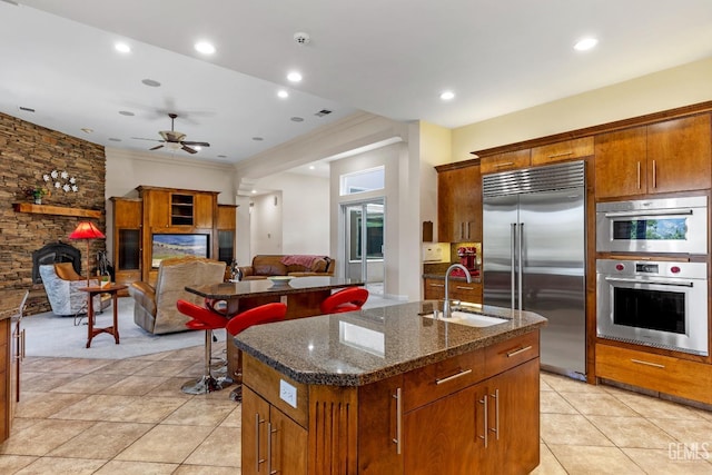 kitchen featuring appliances with stainless steel finishes, ceiling fan, a kitchen island with sink, sink, and a fireplace