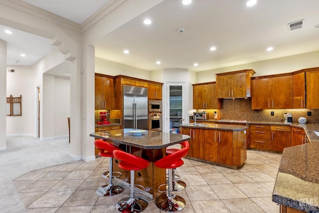 kitchen with stainless steel built in refrigerator, backsplash, dark stone counters, a breakfast bar area, and a kitchen island with sink