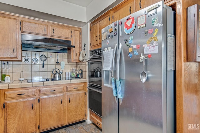 kitchen featuring tasteful backsplash, tile countertops, stone finish flooring, under cabinet range hood, and black appliances