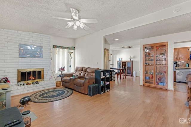 living room featuring a textured ceiling, light wood finished floors, a brick fireplace, and a ceiling fan