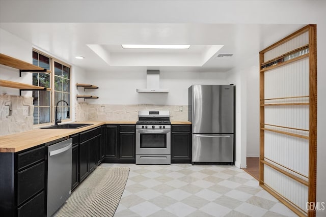 kitchen featuring light floors, open shelves, stainless steel appliances, wall chimney range hood, and dark cabinetry