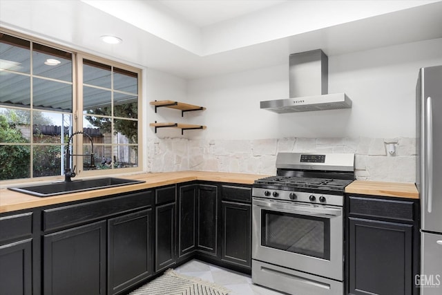 kitchen with stainless steel appliances, dark cabinetry, wall chimney range hood, wooden counters, and a sink