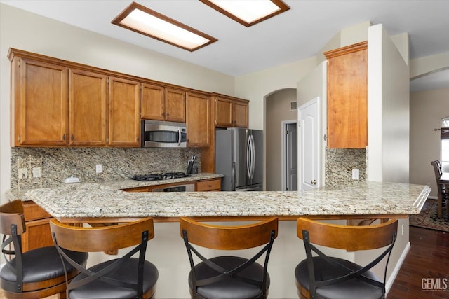 kitchen with appliances with stainless steel finishes, tasteful backsplash, arched walkways, brown cabinets, and a peninsula