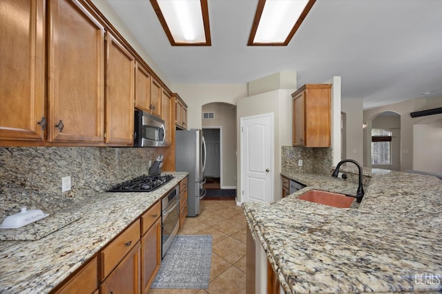 kitchen featuring brown cabinetry, backsplash, appliances with stainless steel finishes, arched walkways, and a sink