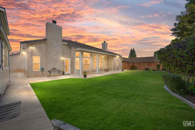 back of house at dusk with a patio, a fenced backyard, stucco siding, a chimney, and a yard