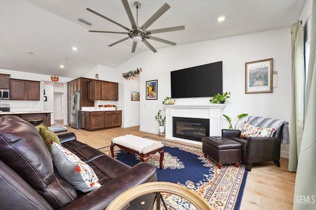 living room with ceiling fan, visible vents, vaulted ceiling, light wood-type flooring, and a glass covered fireplace