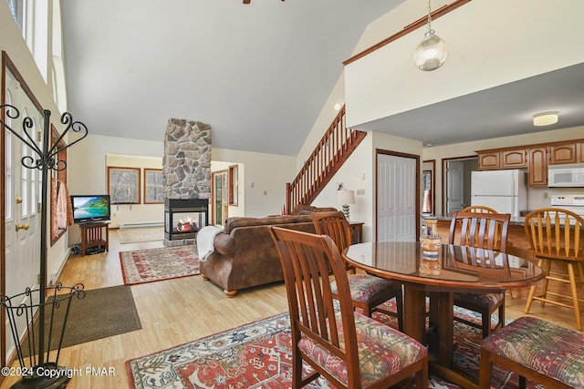 dining area featuring a baseboard radiator, light wood-style flooring, stairway, a fireplace, and high vaulted ceiling