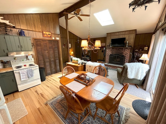 dining area with light wood-style floors, a brick fireplace, wood walls, ceiling fan with notable chandelier, and lofted ceiling with skylight