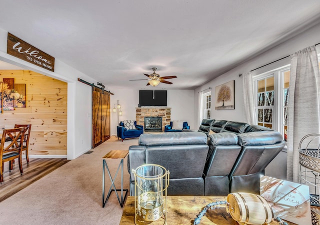 living room with carpet, ceiling fan, a stone fireplace, wood walls, and a barn door