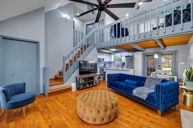 living area with a ceiling fan, a towering ceiling, stairway, light wood-style floors, and beam ceiling