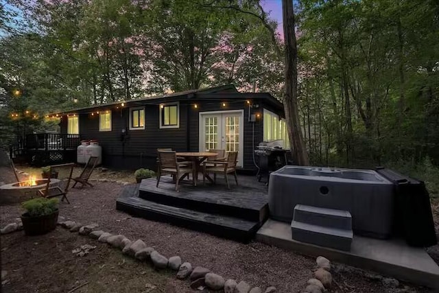 back of house at dusk with a hot tub, a wooden deck, an outdoor fire pit, french doors, and outdoor dining space