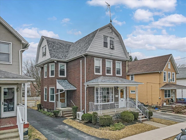 view of front facade with brick siding, a shingled roof, a gambrel roof, a porch, and cooling unit