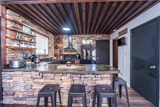 kitchen with wood finished floors, a sink, wall chimney range hood, black appliances, and a wood stove