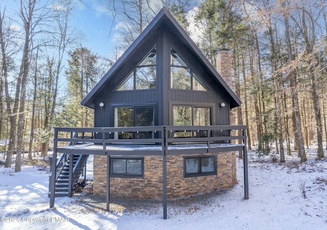 snow covered property with a deck, stone siding, a chimney, and stairs