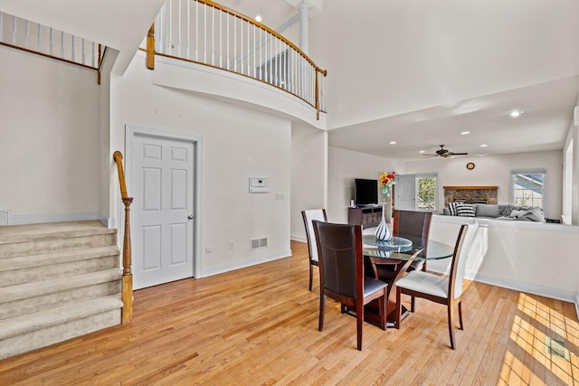 dining space featuring a towering ceiling, light wood-style floors, a stone fireplace, ceiling fan, and stairway