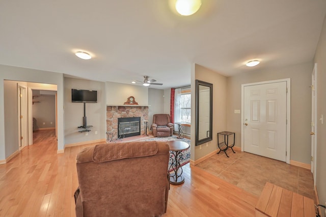 living room with light wood finished floors, a stone fireplace, a ceiling fan, and baseboards