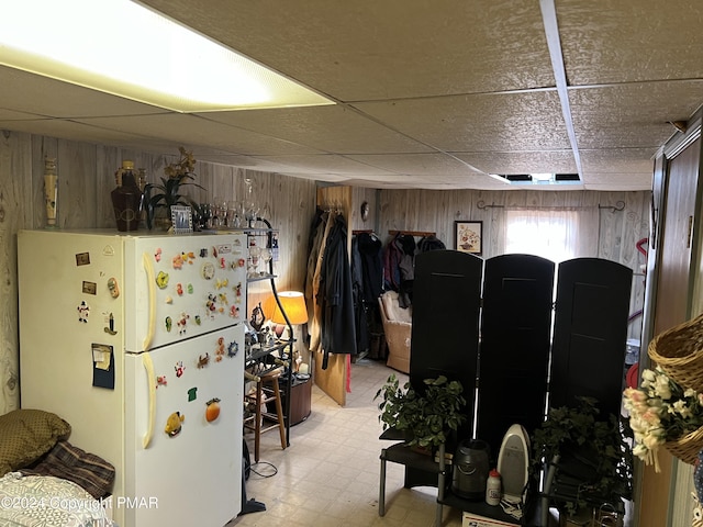 basement with light floors, a drop ceiling, wood walls, and freestanding refrigerator