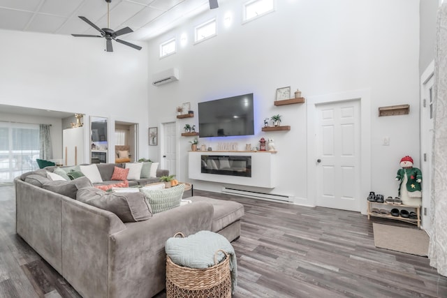 living room featuring a wall unit AC, wood finished floors, a towering ceiling, a ceiling fan, and a glass covered fireplace