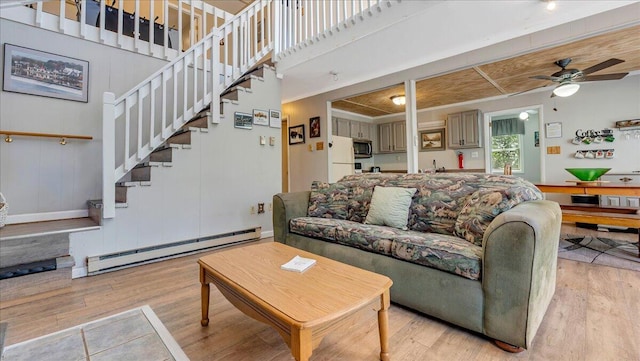 living room featuring a ceiling fan, light wood finished floors, a baseboard heating unit, and stairway