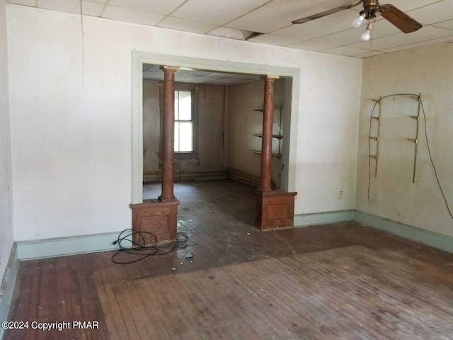 unfurnished room with ornate columns, hardwood / wood-style floors, a ceiling fan, a drop ceiling, and baseboards