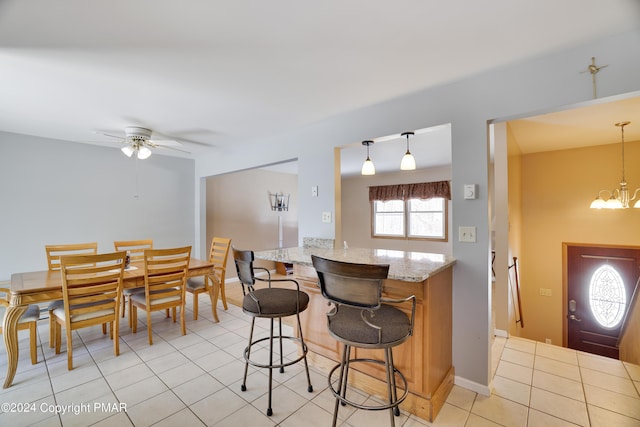 kitchen with light tile patterned floors, light stone counters, ceiling fan with notable chandelier, a kitchen breakfast bar, and pendant lighting