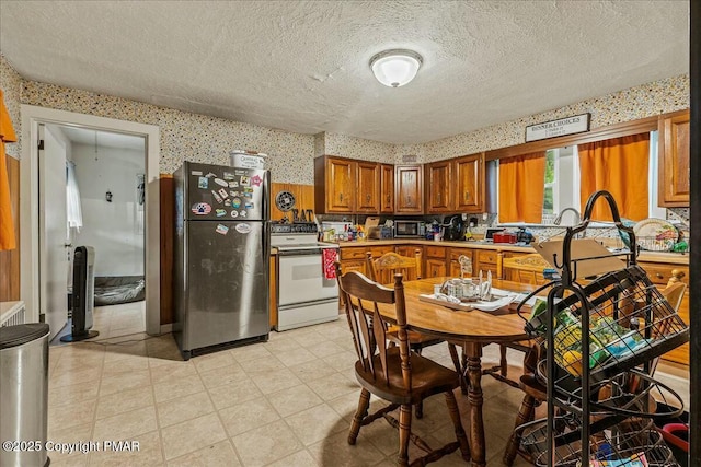 kitchen featuring a textured ceiling, freestanding refrigerator, brown cabinets, white electric range oven, and wallpapered walls